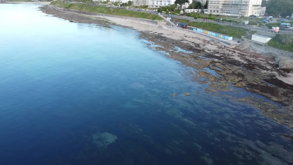 Aerial view of Castle Beach in Falmouth at low tide, showing clear turquoise water, rocky pools, and a row of colorful beach huts.