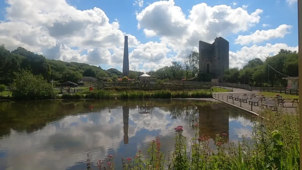 A wide landscape of Lappa Valley featuring a large stone engine house with a tall chimney, a lake with swan-shaped pedalos, and a miniature railway track.